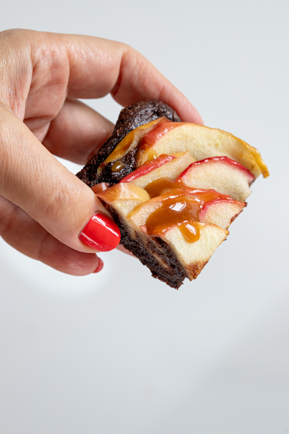A hand with red nail polish holds a fudgy apple cider brownie topped with apple slices, as golden caramel drips down.
