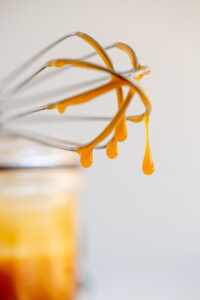 Close-up of a whisk with golden caramel sauce dripping down its wires in thick, glossy streams, warm and shiny against a light background.