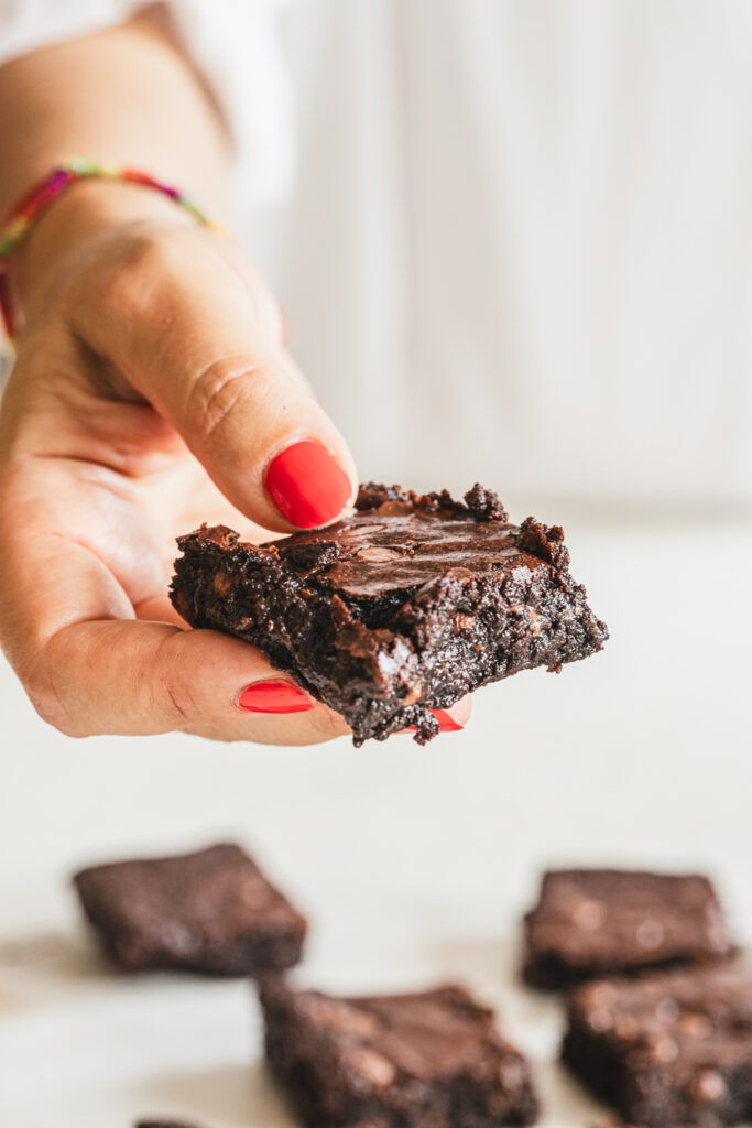 A close-up of a hand with red nail polish holding a shiny, gooey fudgy brownie square, showing its crackly top and rich chocolate texture.