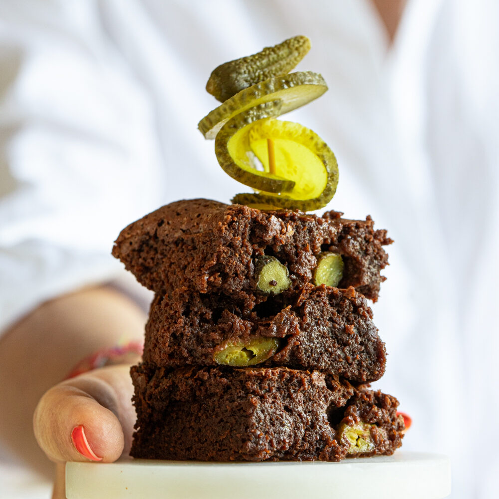 Stack of three Pickle Brownies on a marble coaster, each showing tangy pickle chunks, topped with a skewer of intertwined cucumber slices and a tiny gherkin