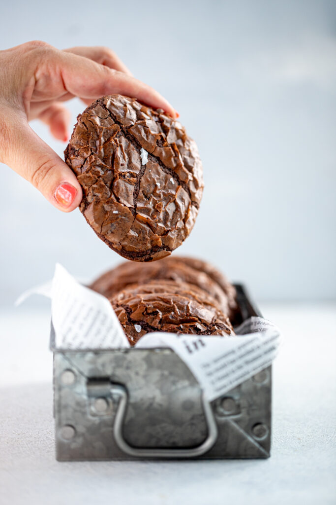 A metal baking pan filled with Salted Chocolate Brownie Cookies, with a hand featuring red nail polish picking one cookie.