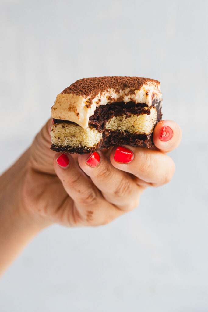 A close-up of a fudgy tiramisu brownie with creamy mascarpone topping, held in a hand with red nail polish. A bite has already been taken, showing the rich, gooey texture of the brownie. Perfect indulgent coffee-flavored dessert.