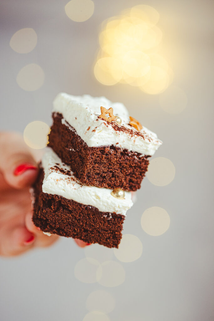 Close-up of a holiday brownie stack, featuring three rich Eggnog Brownie slices with cream cheese frosting, elegantly decorated, in front of a warm, blurry Christmas bokeh.