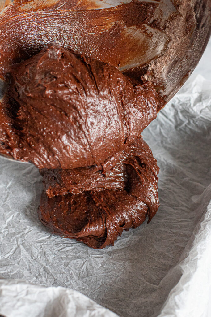Pouring thick, dense brownie batter into a parchment-lined baking pan with a sling for easy removal