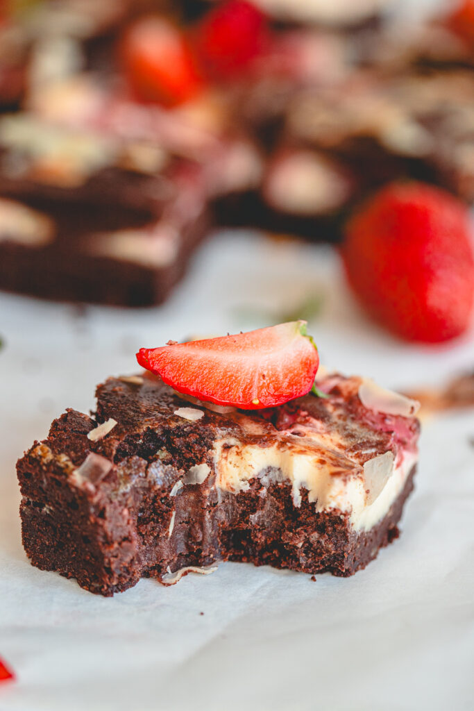 A close-up view of a single fudgy strawberry cheesecake brownie with a bite taken out, revealing the moist chocolate and creamy swirl texture inside, with a blurred row of other brownies lined up on parchment paper in the background.