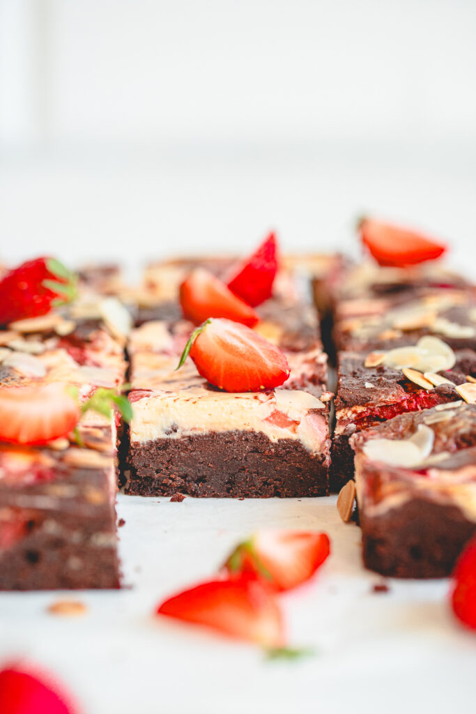 Eye-level photograph of several square-cut flourless strawberry cheesecake brownies arranged on white crumpled parchment paper, showcasing both the deeply fudgy dark chocolate side profile and the toasted sliced almond and baked strawberry topping in natural daylight.