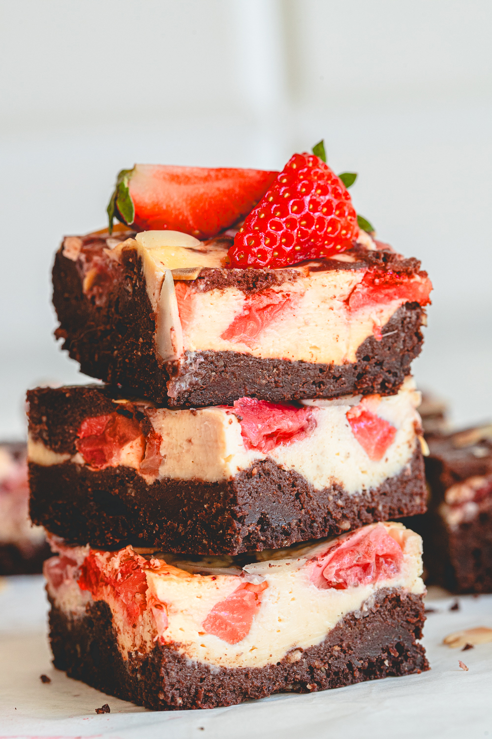 A tall stack of three flourless strawberry cheesecake brownies on white crumpled parchment paper, topped with a fresh halved strawberry, showcasing the contrasting textures of the dense fudgy base, the creamy marble swirl, and the crunchy toasted almonds in sharp detail.