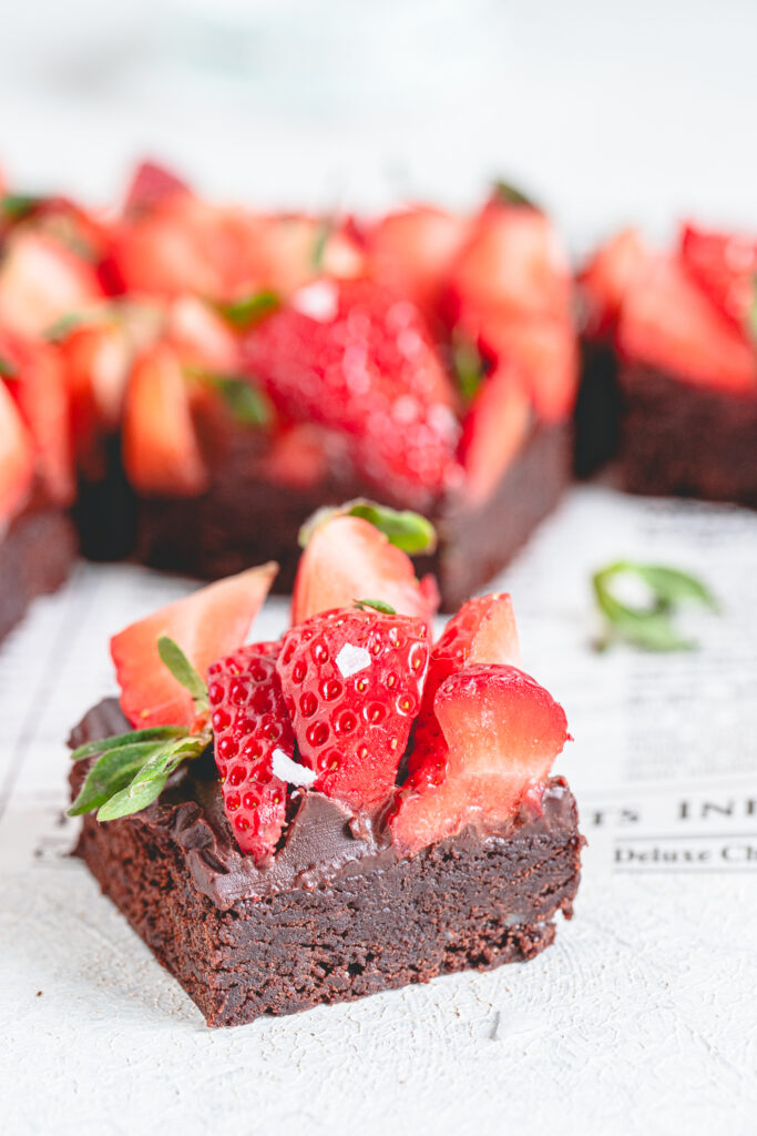 Gourmet fudgy strawberry chocolate brownie square topped with fresh strawberries and flaky sea salt on a vintage newspaper backdrop.
