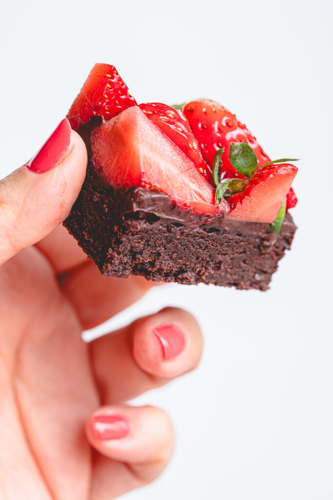 A hand holding a single square of fudgy strawberry chocolate brownie against a clean white background.
