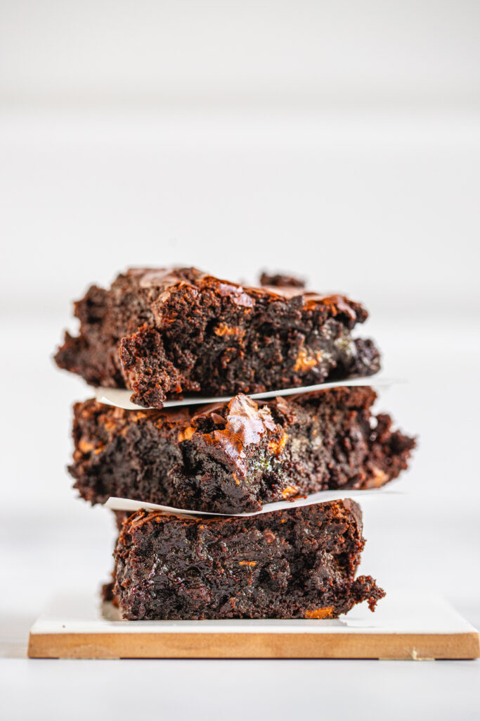 A stack of three rich chocolate brownies sliced while still warm, showing a messy, ultra-fudgy texture with crumbles on a white background.
