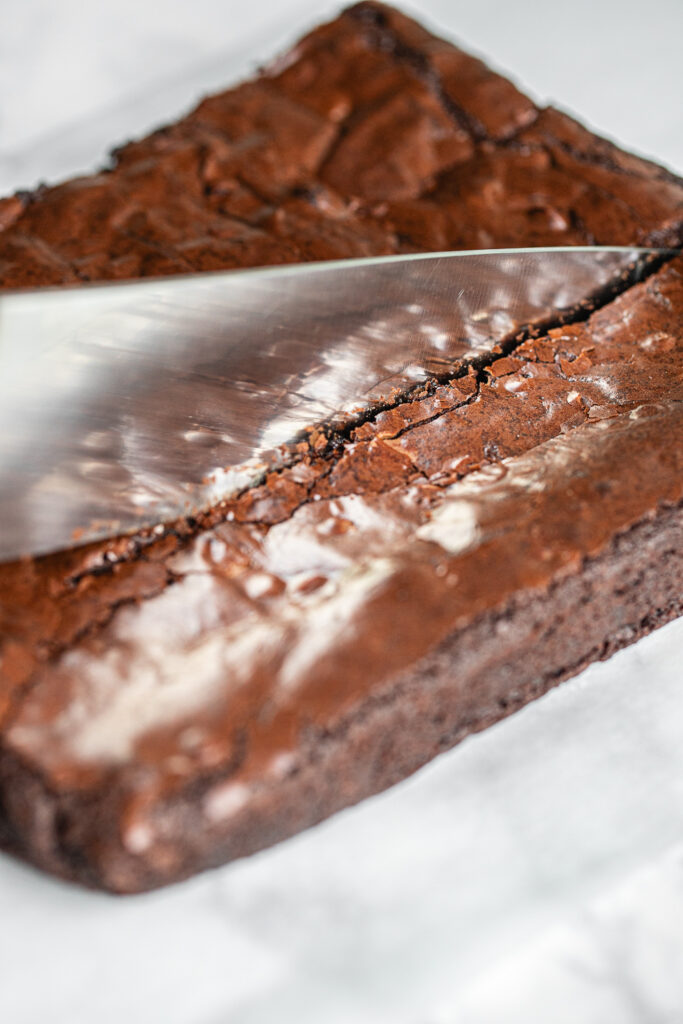 A sharp knife cutting through a chilled brownie slab on parchment paper showing a clean, straight cut