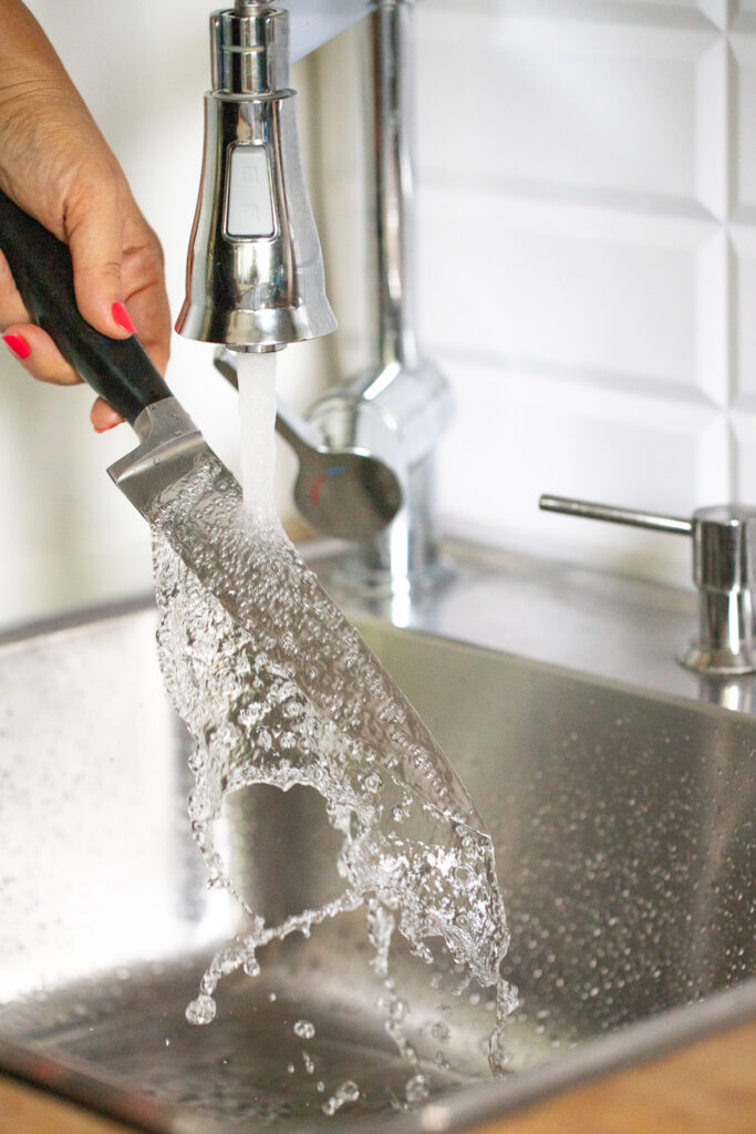 A chef holding a sharp kitchen knife under hot running tap water in a kitchen sink to perform the hot knife trick for cutting brownies.
